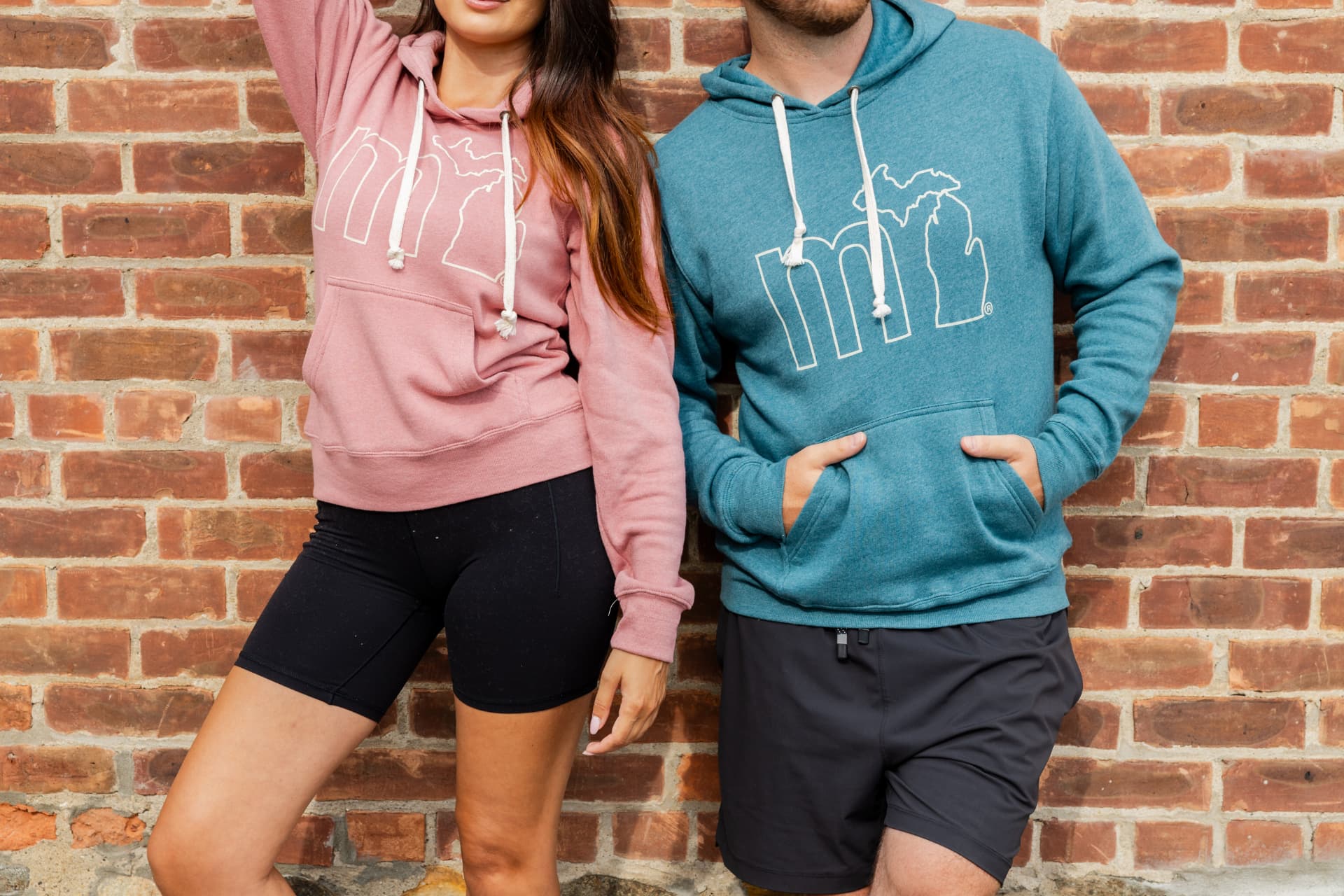 Man and woman wearing teal and rose-pink MI hoodies against a brick wall