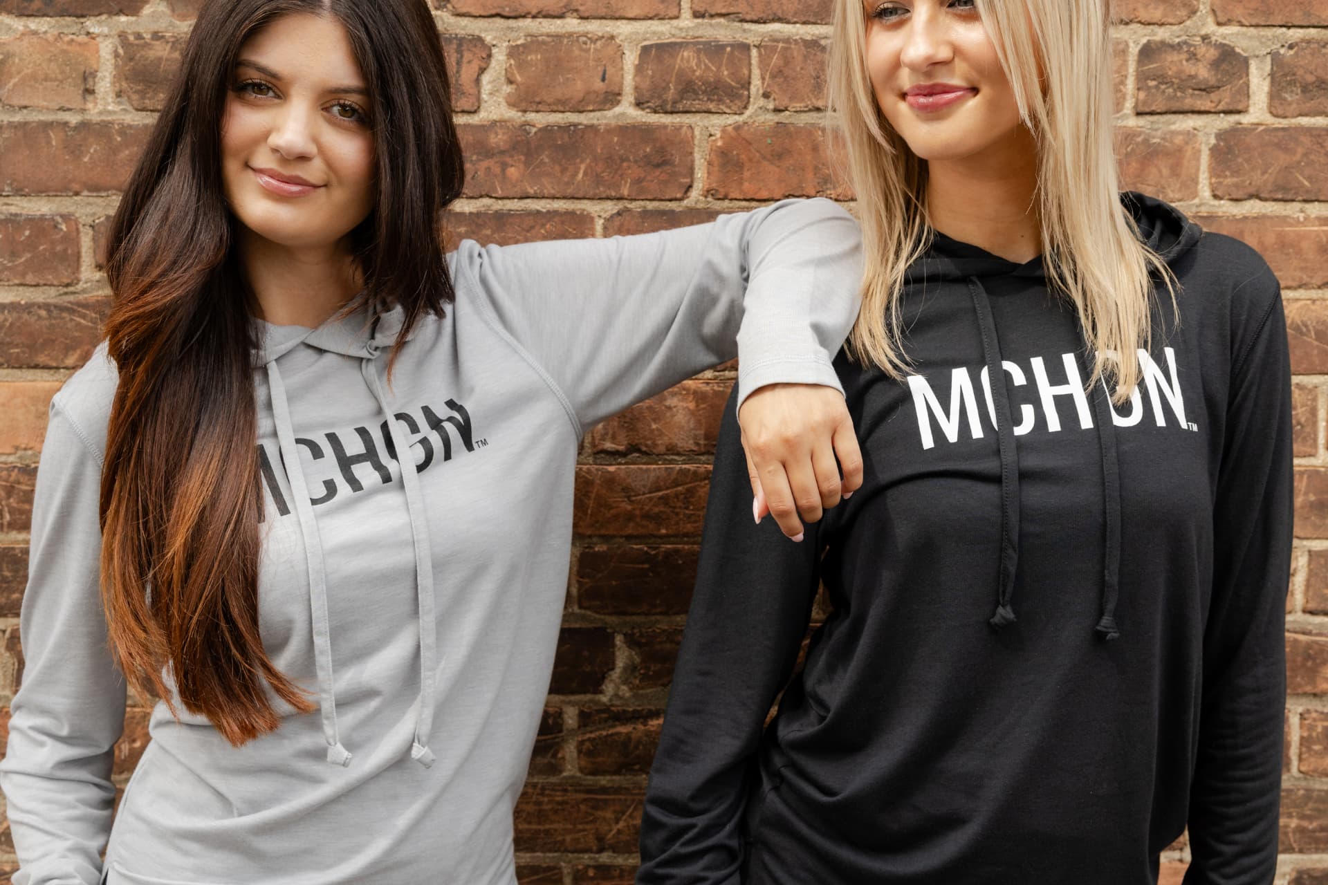 Two women wearing grey and black MCHGN hoodies against a brick wall