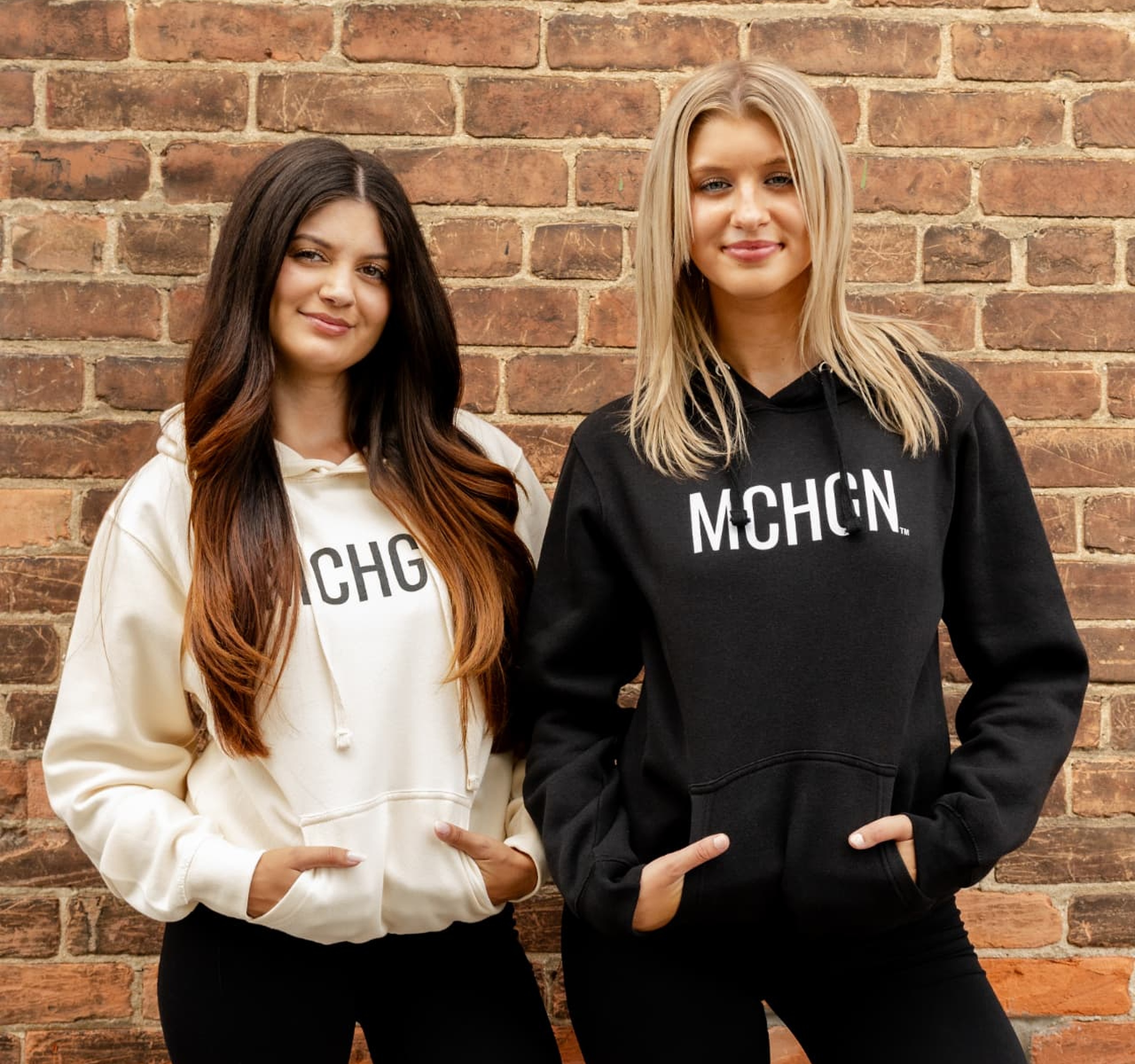 Two women wearing cream and black MCHGN hoodies against a brick wall